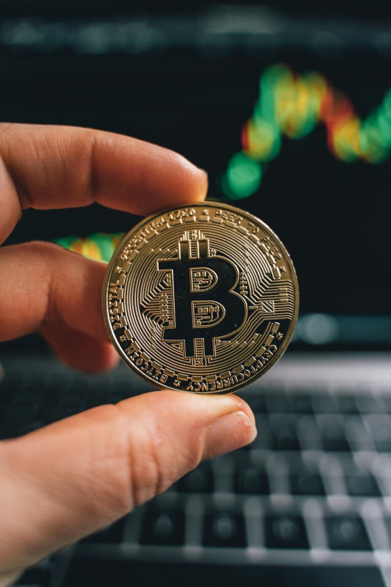 Close-up of a hand holding a Bitcoin coin in front of a trading screen, symbolizing cryptocurrency trading.